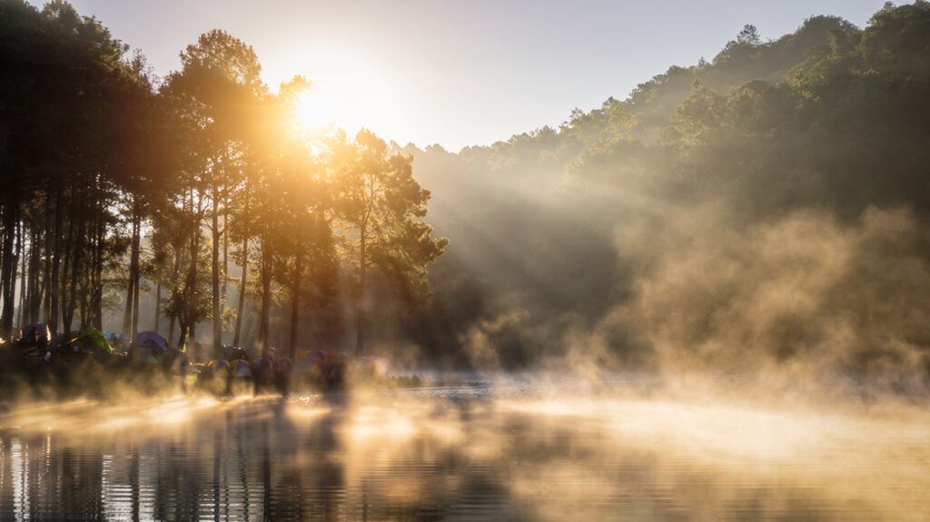 Serene scene with the rising sun over a forested hill by a lake; some tents set up close to the water